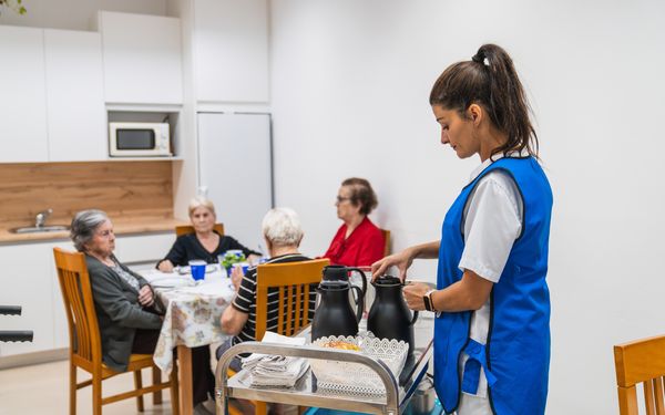 Ein junge Küchenhilfe-Mitarbeiterin bereitet den Tee zum Ausschenken vor. Im Hintergrund sitzen 4 Seniorenheimbewohner an einem Tisch und warten auf den Tee.