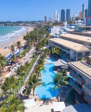 Vista aérea de praia com prédios, piscina, coqueiros e cadeiras de praia azuis à beira-mar em Natal.