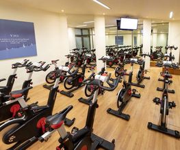 Sala de spinning con bicicletas estáticas, suelo de madera y pantalla en la pared.