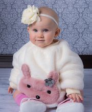 Ein Baby in weißem Outfit mit rosa Häschen, Schleifenstirnband, sitzt auf Holzboden.