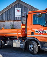 Ein orangefarbener LKW mit der Aufschrift "Fuchs" steht vor einer Holzscheune.