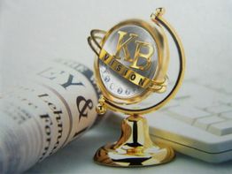 Goldenes KB-Modell auf Ständer, neben Zeitung und Tastatur auf hellem Hintergrund.