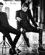 Due uomini in abiti eleganti neri seduti in un ambiente urbano, in bianco e nero.