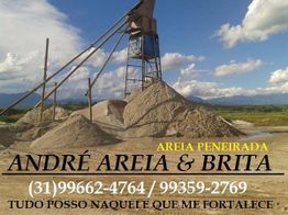 Máquina de peneirar areia com montes de areia e brita ao redor, céu azul e montanhas ao fundo.