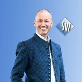 Mann in traditioneller Jacke vor blauem Hintergrund, lächelnd, mit „SW“-Logo rechts oben.