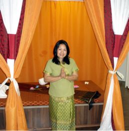 Frau in traditioneller Kleidung steht lächelnd zwischen Vorhängen mit gefalteten Händen.