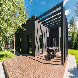Modern black house with pergola and wooden deck, surrounded by trees and greenery.