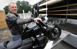 Mann lädt Motorrad auf Anhänger, trägt eine Lederjacke, umgeben von Baum und Anhänger.