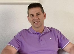 Ein Mann in einem lila Polohemd sitzt lächelnd auf einem Sessel vor einer weißen Wand.