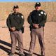 Two uniformed officers standing on a sandy outdoor area under a clear blue sky.