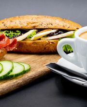 Brötchen mit Wurst und Käse, daneben eine Cappuccino-Tasse auf einem Tablett, Gurkenscheiben vorne.