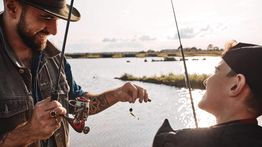 Two people smiling while fishing by a lake on a sunny day.