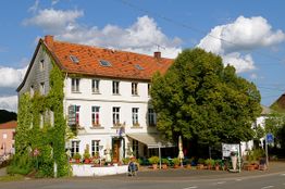 Weißes, begrüntes Gebäude an einer Ecke, umgeben von Bäumen, mit blauen Himmel und Wolken.