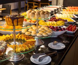 Buffet de café da manhã com pães, bolos e frutas em mesas de vidro.