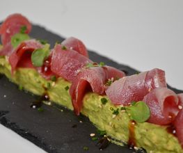 Plato de atún rojo sobre aguacate con sésamo y aderezo en bandeja oscura.