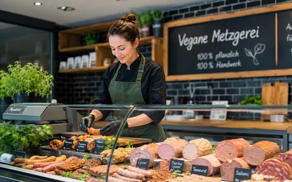Moderne vegane Metzgerei mit Verkaufstheke, pflanzlichen Wurst- und Fleischalternativen sowie einer Mitarbeiterin bei der Präsentation handwerklich hergestellter Produkte.