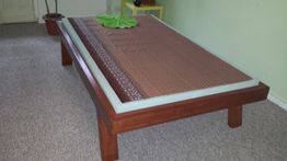 Holzbett mit brauner Decke, grünes Kissen, Topfpflanze und Tisch im Hintergrund.