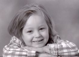 Ein Kind mit kariertem Hemd lächelt und stützt den Kopf auf die Arme, Schwarz-Weiß-Foto.