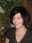 Eine lächelnde Person mit kurzen, dunklen Haaren sitzt vor einer Pflanze.