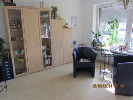 Helles Zimmer mit Bücherschrank, Pflanzen, zwei blauen Stühlen und einem Tisch am Fenster.