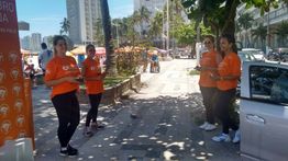 Mulheres de camiseta laranja em calçadão ensolarado, próximas a prédios e árvores.