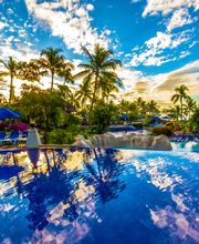 Piscina rodeada de palmeras y tumbonas azules, cielo azul con nubes blancas reflejado en el agua.