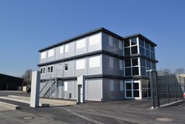 Graues, dreistöckiges Containergebäude mit Treppe und Zaun bei sonnigem Wetter.