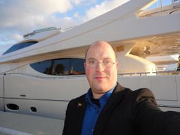 Eine Person macht ein Selfie vor einer großen weißen Jacht bei sonnigem Wetter.