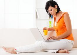 Frau in orangener Jacke arbeitet am Laptop und hält ein Glas Saft auf einer Couch sitzend.