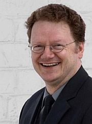 Eine lächelnde Person mit Brille und Anzug steht vor einer weißen Backsteinwand.