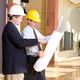 Two men in hard hats review architectural blueprints in a bright, modern lobby.