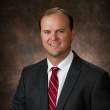 Man in a suit with a red tie, smiling, brown background.