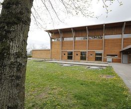 Holzgebäude neben grünem Rasen und Baum im Vordergrund. Überdachter Bereich rechts.