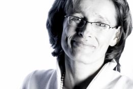 Eine Person mit Brille und hellem Pullover lächelt vor einem weißen Hintergrund.