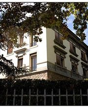 Weiße Villa hinter einem schmiedeeisernen Zaun, umgeben von Bäumen und Sonnenlicht.