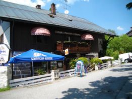 Rustikales Café mit Terrasse, blauem Sonnenschirm und Schildern, ländliche Umgebung, sonniger Tag.