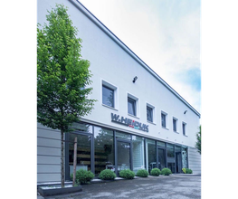 Modernes Bürogebäude mit großen Fenstern, davor ein Baum und Büsche, blauer Himmel.
