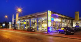 Autohaus bei Nacht mit beleuchteten Schildern und parkenden Autos davor.