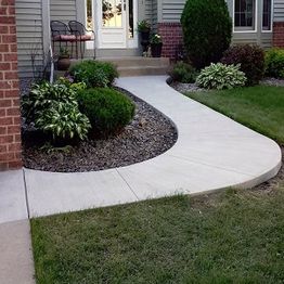 Curved walkway with bushes and plants leading to a house entrance with chairs.