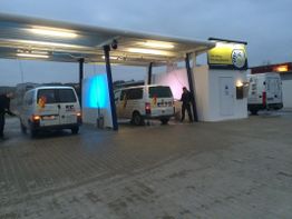 Mehrere weiße Lieferwagen an einer Autowaschanlage, Männer waschen Fahrzeuge bei Dämmerung.