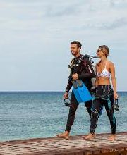 Pareja caminando en muelle con equipo de buceo frente al mar turquesa.