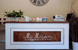 Bancone in legno bianco con decorazioni, orologio grande e piante sopra, insegna "Ma Maison".