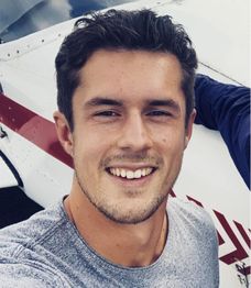 Junger Mann lächelt vor einem Flugzeug in die Kamera.