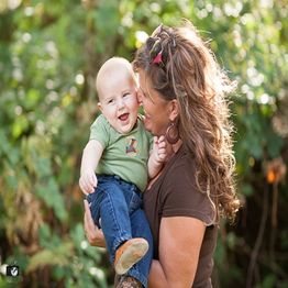 Woman holding and smiling at a laughing baby outdoors with greenery in the background.