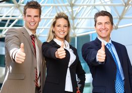 Drei Personen in Anzügen zeigen Daumen hoch und lächeln, im Hintergrund ein modernes Gebäude.