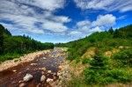 Ein ruhiger Fluss fließt durch eine grüne, hügelige Landschaft unter einem blauen Himmel mit Wolken.