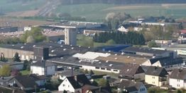 Luftaufnahme eines Gewerbegebiets und Wohnhäusern vor einem grünen, hügeligen Hintergrund.