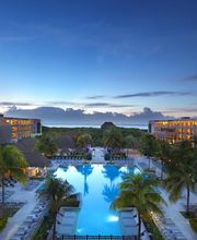 Complejo turístico con piscina al atardecer, rodeado de palmeras y edificios iluminados.