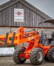 Orange Radlader vor einer Holzscheune mit mehreren Schildern, grauer Himmel im Hintergrund.