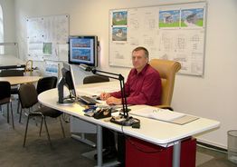 Mann im Büro am Schreibtisch, umgeben von Architekturplänen, arbeitet am Computer.